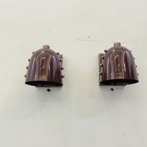 Op de afbeelding: Twee bijpassende wandkandelaars met een klokvormig ontwerp. De wandkandelaars hebben een diepe bordeauxrode kleur met een metallic afwerking en zijn aan een muur bevestigd. Elke wandkandelaar heeft een reeks kleine, ronde klinknagels rond de basis van de klok.