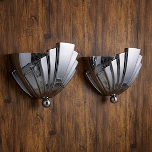 Puede incluir: Dos apliques de pared plateados pulidos con un diseño en forma de abanico, montados sobre una superficie de madera. Cada aplique tiene una base redondeada y una serie de elementos rectangulares radiantes. Los apliques reflejan el entorno.
