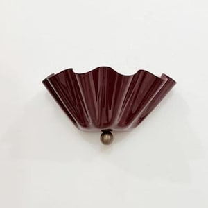 Op de afbeelding: Bordeaux wandlamp met een gerimpeld, schelpachtig ontwerp. De lamp heeft een glanzende afwerking en een klein, rond, messingkleurig accent aan de onderkant. De armatuur is op een witte muur gemonteerd.
