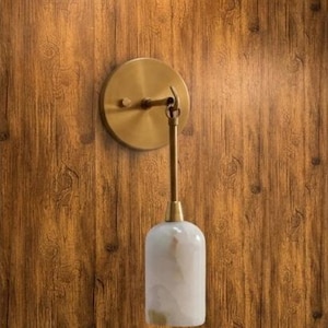 Può includere: Applique da parete moderna con finitura in ottone, con una piastra rotonda e un braccio curvo che supporta un paralume bianco e beige effetto marmo. L'applique è montata su una parete di legno.