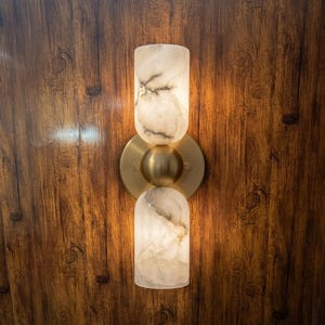 May include: A modern wall sconce with a brass finish and two alabaster glass shades. The shades are cylindrical and have a marbled pattern. The sconce is mounted on a wooden wall.