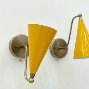 Op de afbeelding: Twee wandkandelaars met heldergele conische kappen en messingkleurige hardware. De kappen zijn schuin en de wandkandelaars zijn op ronde achterplaten gemonteerd. Het ontwerp is modern.