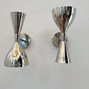 May include: Two polished chrome wall sconces with a mid-century modern design. Each sconce features a flared top and bottom, connected by a narrower center. Small circular perforations adorn the top rim of each sconce. The sconces are mounted on circular backplates.