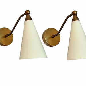 Könnte beinhalten: Zwei Wandleuchten mit Messing-Finish. Jede Leuchte hat einen kreisförmigen Sockel, einen gebogenen Arm und einen konischen, cremefarbenen Schirm. Das Design ist minimalistisch und modern, geeignet für die Innenbeleuchtung.