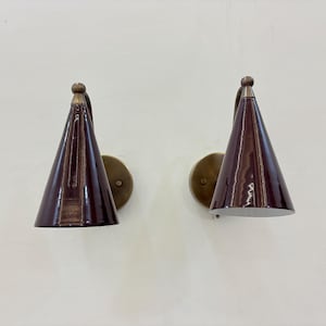Può includere: Due applique da parete con paralumi conici bordeaux scuro e basi circolari color ottone. Le applique sono montate su una parete bianca. Presentano un design in stile moderno della metà del secolo.