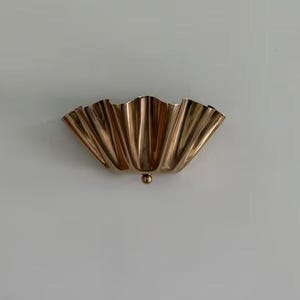 May include: A gold-colored, wall-mounted sconce with a scalloped, fan-like design. The sconce has a small, round detail at the bottom. The fixture is made of metal and is mounted on a white wall. This decorative piece is suitable for home decor.