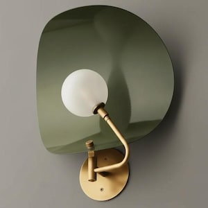 Könnte beinhalten: Moderne Wandleuchte mit einem großen, olivgrünen, asymmetrischen Schirm. Die Leuchte hat eine weiße Kugellampe und einen gebogenen, messingfarbenen Arm. Der Sockel ist eine runde, messingfarbene Platte.