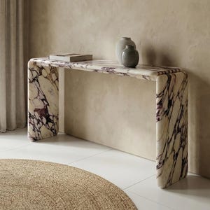 Puede incluir: Una consola moderna de mármol con un diseño curvo. La mesa presenta un patrón veteado blanco y morado. Dos jarrones grises y un libro descansan sobre la superficie de la mesa. La mesa está colocada contra una pared de tonos neutros y una ventana con una cortina transparente.