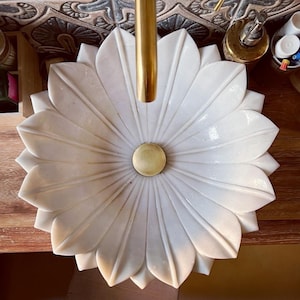 Op de afbeelding: Witte, bloemvormige marmeren wastafel met een gouden kraan. De wastafel heeft een centrale gouden afvoerafdekking en staat op een houten oppervlak. Het ontwerp van de wastafel lijkt op een bloeiende bloem.