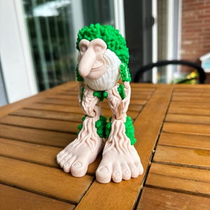May include: A quirky figurine with a prominent nose, green hair, and tree-like limbs and feet. The figure is mainly off-white with green details, positioned on a wooden table. The design evokes a forest creature.