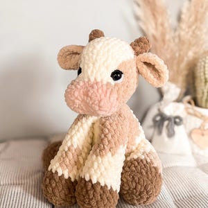 K&ouml;nnte beinhalten: Ein handgefertigtes H&auml;keltier in Form einer Kuh in Creme-, Hellbraun- und Dunkelbraunt&ouml;nen. Das Stofftier hat eine rosa Schnauze und schwarze Sicherheitsaugen. Die Kuh sitzt auf einer gestreiften Oberfl&auml;che.
