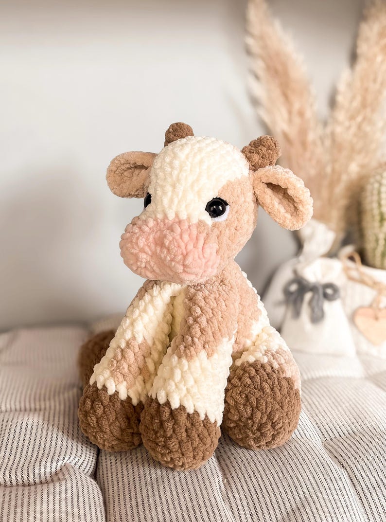 K&ouml;nnte beinhalten: Ein handgefertigtes H&auml;keltier in Form einer Kuh in Creme-, Hellbraun- und Dunkelbraunt&ouml;nen. Das Stofftier hat eine rosa Schnauze und schwarze Sicherheitsaugen. Die Kuh sitzt auf einer gestreiften Oberfl&auml;che.