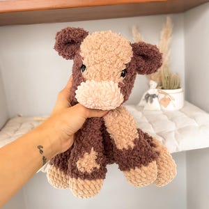 K&ouml;nnte beinhalten: Ein handgefertigtes Kuscheltier in Form einer Kuh, in Braun- und Beiget&ouml;nen. Das weiche, strukturierte Spielzeug hat ein hellbeige Gesicht und Flecken, mit dunkelbraunen Akzenten an Ohren, Beinen und K&ouml;rper. Die Kuh wird vor einem Regal gehalten.