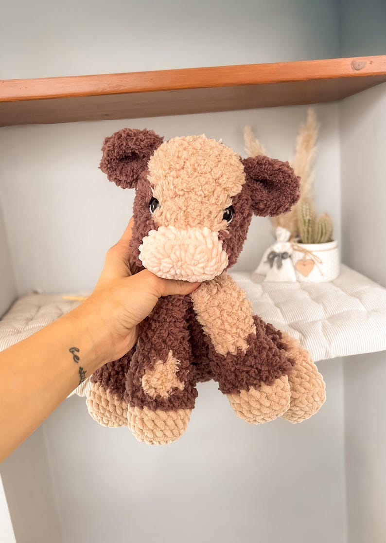 K&ouml;nnte beinhalten: Ein handgefertigtes Kuscheltier in Form einer Kuh, in Braun- und Beiget&ouml;nen. Das weiche, strukturierte Spielzeug hat ein hellbeige Gesicht und Flecken, mit dunkelbraunen Akzenten an Ohren, Beinen und K&ouml;rper. Die Kuh wird vor einem Regal gehalten.