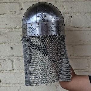May include: A silver-colored, medieval-style helmet with intricate etched designs and a chainmail face guard. The helmet has a rounded top and is secured with rivets. The chainmail hangs down, providing facial protection.