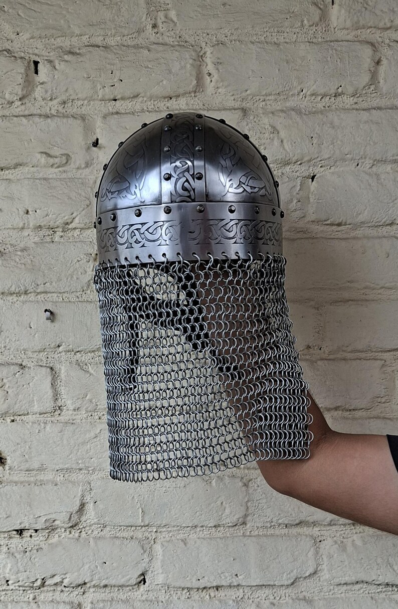 May include: A silver-colored, medieval-style helmet with intricate etched designs and a chainmail face guard. The helmet has a rounded top and is secured with rivets. The chainmail hangs down, providing facial protection.