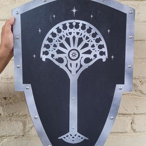 Könnte beinhalten: Ein schwarzes Schild mit silberner Umrandung und einem stilisierten weißen Baumdesign. Das Schild hat ein Sternmuster und wird vor einer Backsteinmauer gehalten. Das Schild ist etwa 61 cm hoch.