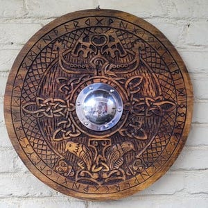 Peut inclure: Un bouclier rond en bois avec des gravures complexes de motifs de l'époque viking. Le bouclier présente un bossage central en métal poli et est monté sur un mur de briques blanches. Les gravures incluent des navires vikings et des runes.