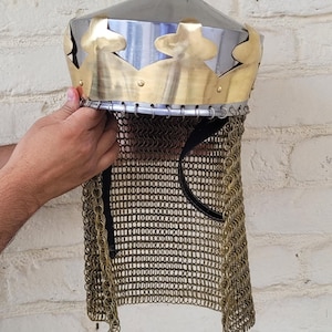 Könnte beinhalten: Ein mittelalterlicher Helm mit silberner konischer Spitze und einem goldfarbenen, kronenförmigen Rand. Der Helm hat einen Kettenkragen und wird vor einer weißen Backsteinmauer gehalten.