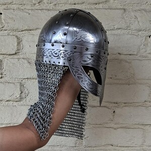 May include: A silver-colored Viking helmet with intricate engraved patterns and a chainmail neck guard. The helmet features a rounded top with rivets and a decorative visor. The design includes Celtic knotwork.