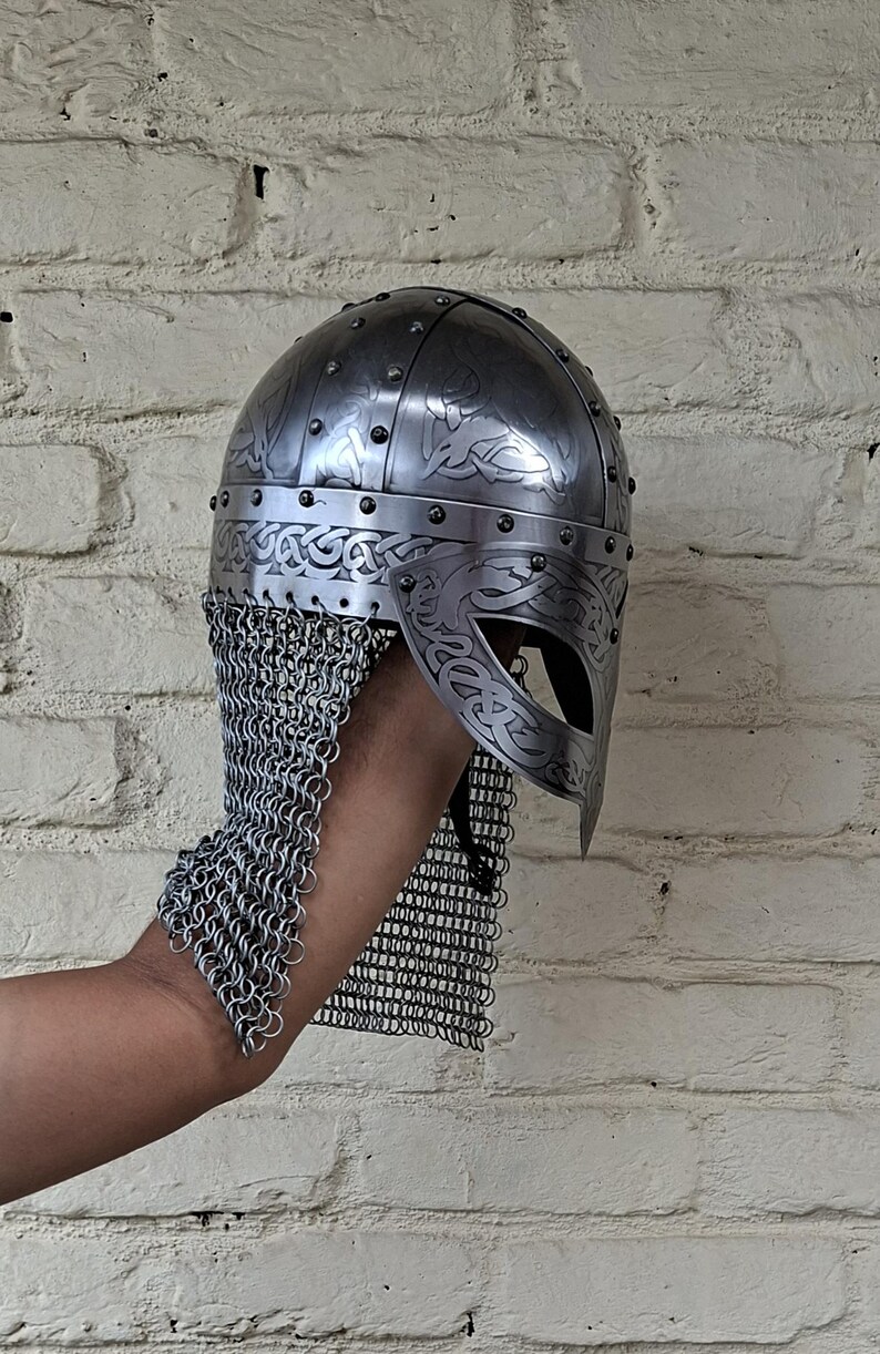 May include: A silver-colored Viking helmet with intricate engraved patterns and a chainmail neck guard. The helmet features a rounded top with rivets and a decorative visor. The design includes Celtic knotwork.
