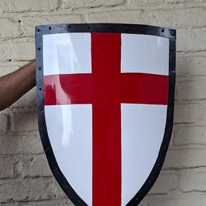 Puede incluir: Un escudo con fondo blanco y una gran cruz roja en el centro. El escudo tiene un borde negro con remaches. El escudo se sostiene contra una pared de ladrillos.