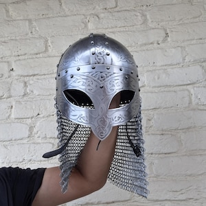 May include: A silver-colored Viking helmet with intricate engraved patterns and eye openings. The helmet features a chainmail neck guard. The design includes decorative rivets and a detailed faceplate.