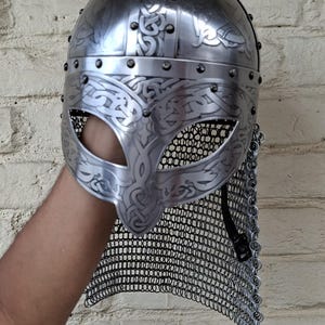 May include: A silver-colored Viking helmet with intricate engraved patterns and a chainmail neck guard. The helmet features a rounded top and a face guard with eye openings. The design includes decorative rivets.