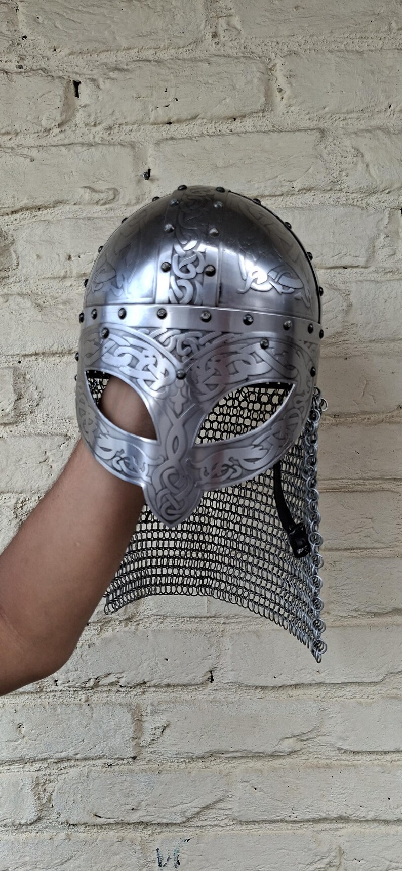 May include: A silver-colored Viking helmet with intricate engraved patterns and a chainmail neck guard. The helmet features a rounded top and a face guard with eye openings. The design includes decorative rivets.