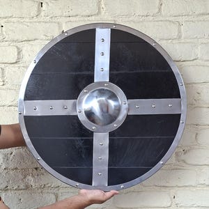 Op de afbeelding: Ronde, zwarte houten schild met een zilveren metalen rand en kruisontwerp. Het schild heeft een centrale, gepolijste metalen knop. Gehouden tegen een witte bakstenen muur.