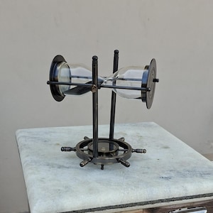 May include: An hourglass with a dark metal frame and a ship's wheel base. The glass bulbs hold black sand. The hourglass is supported by two vertical rods. The base is a ship's wheel design with spokes. The hourglass is on a marble-topped table.
