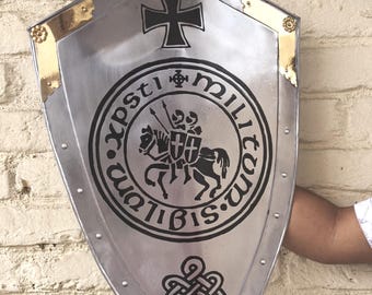 May include: A silver-colored shield with a black cross at the top and a circular emblem featuring two knights on horseback. The shield has gold-colored accents and rivets along the edges. The shield is held against a white brick wall.