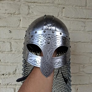 May include: A silver-colored Viking helmet with intricate engraved patterns and a chainmail neck guard. The helmet features a rounded top, eye openings, and decorative rivets. The design is reminiscent of historical armor.