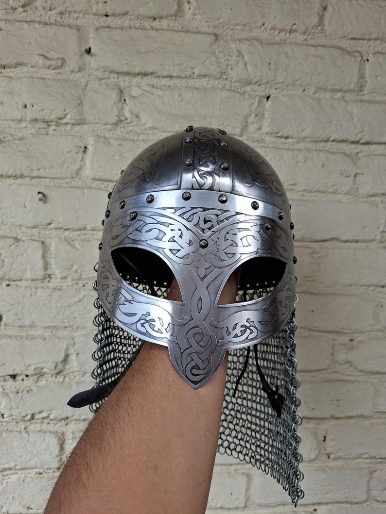 May include: A silver-colored Viking helmet with intricate engraved patterns and a chainmail neck guard. The helmet features a rounded top, eye openings, and decorative rivets. The design is reminiscent of historical armor.