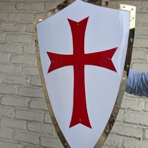 Puede incluir: Un escudo blanco con un emblema de cruz roja, enmarcado por un borde dorado. El escudo se sostiene contra una pared de ladrillos. La cruz tiene una forma distintiva, y el borde está asegurado con remaches.