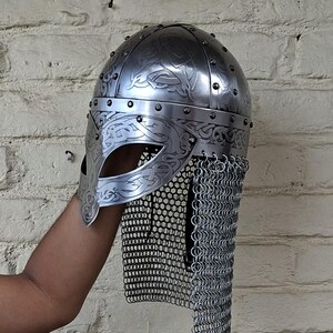 May include: A silver-colored Viking helmet with intricate engraved patterns and a chainmail neck guard. The helmet features a curved visor and is riveted together. The design includes decorative swirls and lines, typical of Viking artistry.