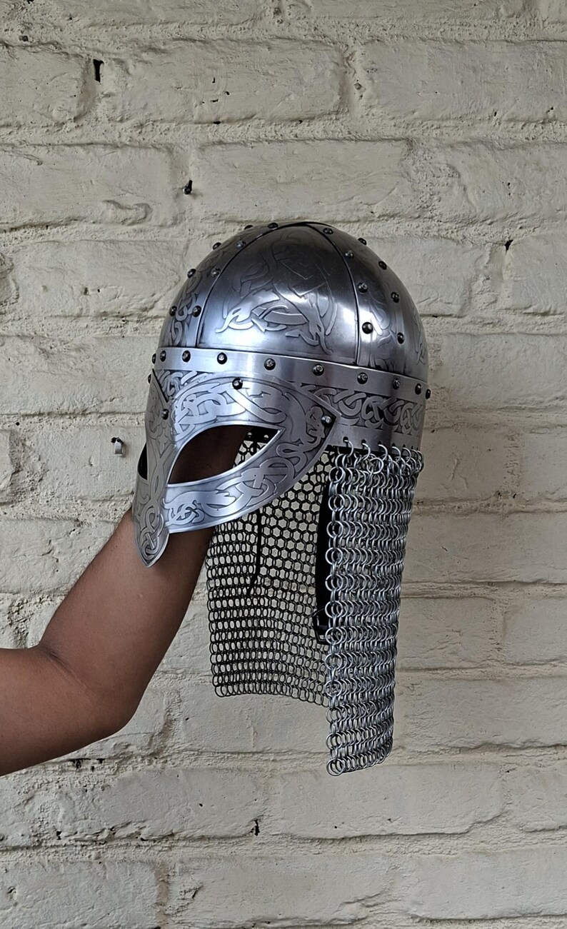 May include: A silver-colored Viking helmet with intricate engraved patterns and a chainmail neck guard. The helmet features a curved visor and is riveted together. The design includes decorative swirls and lines, typical of Viking artistry.