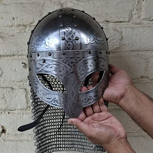 May include: A silver-colored Viking helmet with intricate engraved designs and a chainmail neck guard. The helmet features decorative patterns and rivets, showcasing a historical aesthetic. The helmet is held against a white brick wall.