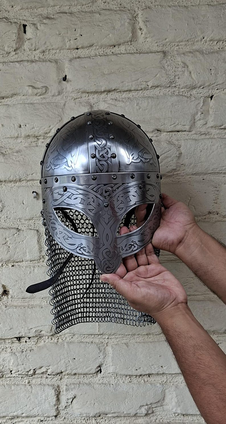 May include: A silver-colored Viking helmet with intricate engraved designs and a chainmail neck guard. The helmet features decorative patterns and rivets, showcasing a historical aesthetic. The helmet is held against a white brick wall.