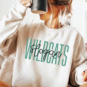 May include: A cream-colored crewneck sweatshirt featuring the word WILDCATS printed in large, vertical, teal sans-serif letters, with the word slogan written in a black, cursive script font layered horizontally across the center of the text.