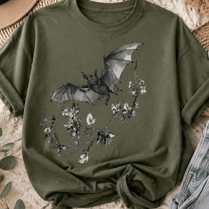 May include: An olive green short-sleeve t-shirt featuring a black and white graphic print of a bat in flight above a flowering branch. The crew neck shirt has a relaxed fit with rolled sleeves and a detailed illustration of nature.