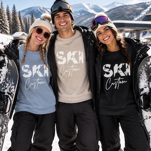 May include: Three crewneck sweatshirts in light blue, beige, and black, each featuring the white text 'SKI' with a mountain graphic inside the letters and the word 'Custom' in a cursive script font underneath, worn with black ski pants and jackets.