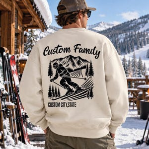 May include: A cream-colored crewneck sweatshirt featuring a black graphic on the back. The design depicts a skier on a mountain with pine trees and the text Custom Family above and CUSTOM CITY,STATE below the illustration.