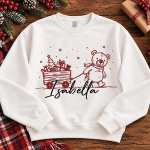 以下が含まれることがあります： 長袖の白いスウェットシャツ。クリスマスプレゼントとツリーを載せた木製のワゴンを引くテディベアの濃い赤色のグラフィックが描かれています。グラフィックの下には、黒い筆記体でIsabellaという名前がプリントされています。