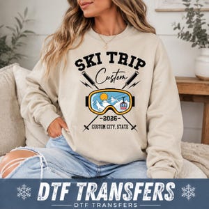 May include: A beige crewneck sweatshirt featuring a graphic print with the text "SKI TRIP", "Custom", "2026", and "CUSTOM CITY, STATE". The design includes black ski poles, a yellow-rimmed ski goggle, a sun, mountains, and a red cable car.