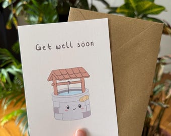 May include: A white greeting card with the text "Get well soon" and a cartoon well illustration. The well has a bandage and a smiling face. A brown envelope is behind the card.