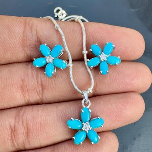 May include: A silver necklace and earring set with a floral design. Each flower has five turquoise-coloured petals and a small, clear stone in the centre. The necklace pendant and earrings are displayed on a hand.