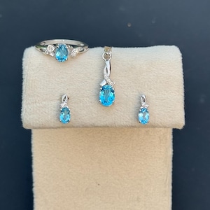 Puede incluir: Un conjunto de joyas con un anillo, colgante y pendientes de plata, todos adornados con piedras preciosas ovales de color azul claro. El anillo tiene un diseño delicado con pequeñas piedras de acento. El colgante y los pendientes reflejan el diseño del anillo.