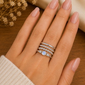Op de afbeelding: Een stapel van vier zilverkleurige ringen gedragen aan een vinger. De set bevat een gladde band, een gedraaide touw-stijl band, een kralenband en een centrale ring met een ronde iriserende maansteen geflankeerd door twee kleine heldere stenen.