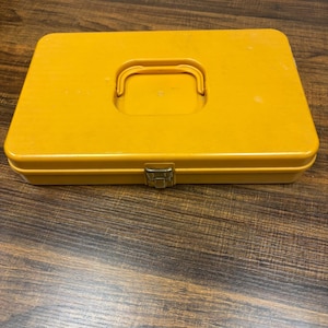 Può includere: Una valigetta rettangolare in plastica gialla dotata di una maniglia incassata sulla superficie superiore e di una singola chiusura a scatto in metallo sul bordo anteriore. La custodia presenta una finitura liscia e un design piatto.